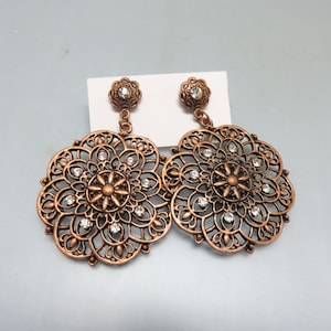 May include: A pair of large, round, copper-colored earrings with intricate filigree designs and clear rhinestones.
