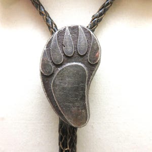 May include: A silver-toned bolo tie featuring a bear paw design. The pendant is attached to a braided black leather cord. The bear paw has five distinct toe pads and a textured surface.