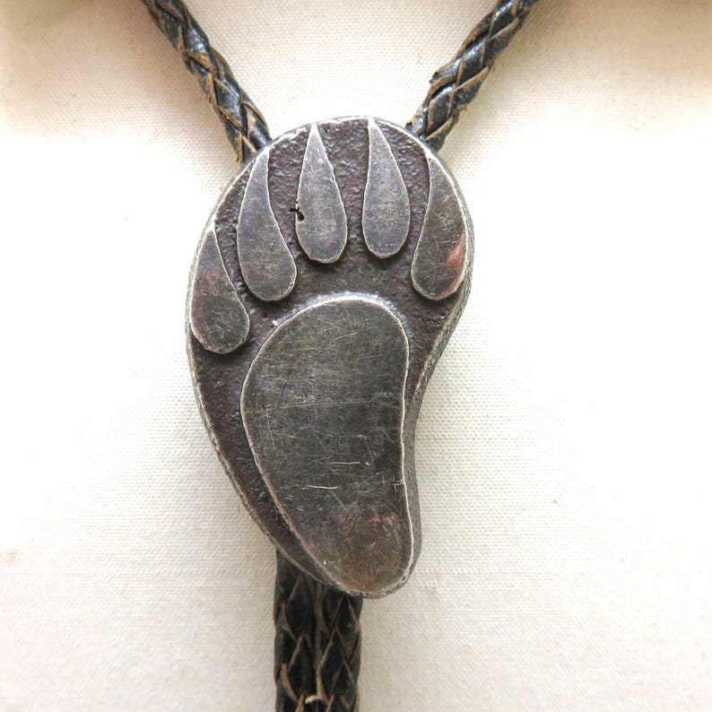 May include: A silver-toned bolo tie featuring a bear paw design. The pendant is attached to a braided black leather cord. The bear paw has five distinct toe pads and a textured surface.