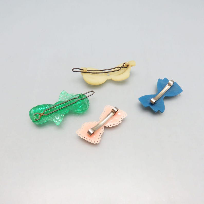 May include: Four vintage hair barrettes in various colors: green, yellow, pink, and blue. The barrettes are shaped like bows and have metal clasps. The green barrette has a textured, fish-like design. The pink barrette has a scalloped edge.