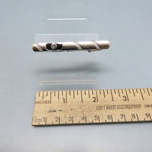 May include: A silver tie clip with a black enamel band and a clear rhinestone. The tie clip is about 1 1/2 inches long.