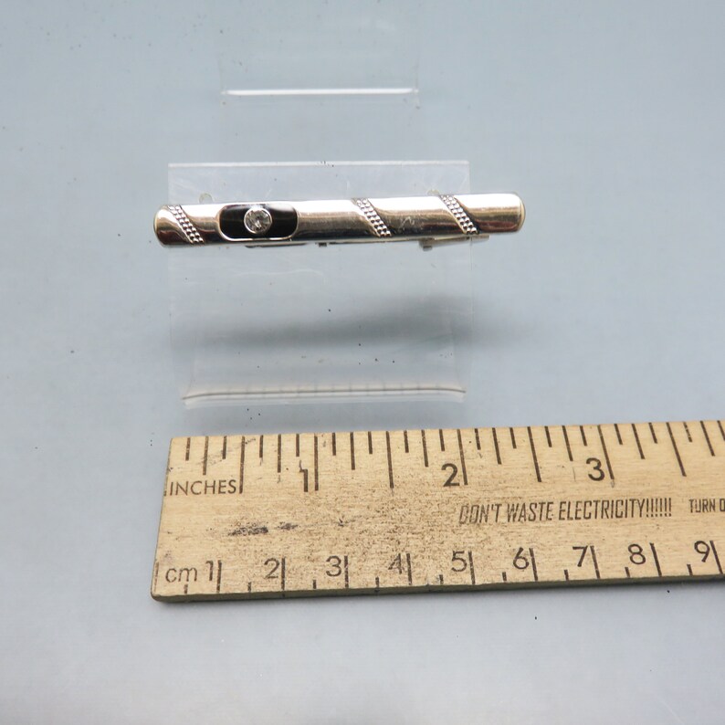 May include: A silver tie clip with a black enamel band and a clear rhinestone. The tie clip is about 1 1/2 inches long.