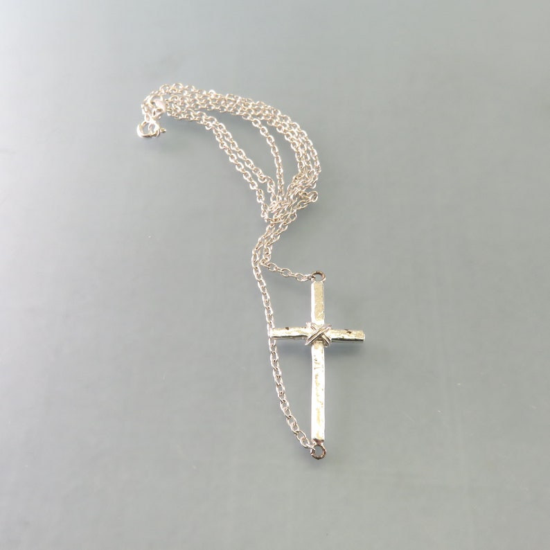K&ouml;nnte beinhalten: Eine silberne Kettenkette mit einem silbernen Kreuzanh&auml;nger. Das Kreuz hat eine strukturierte Oberfl&auml;che und ein gedrehtes Design in der Mitte.