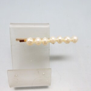 May include: A gold-toned hair barrette featuring a row of seven off-white, round pearls. The barrette is displayed on a clear, rectangular stand against a white background. This elegant hair accessory is suitable for various hairstyles.