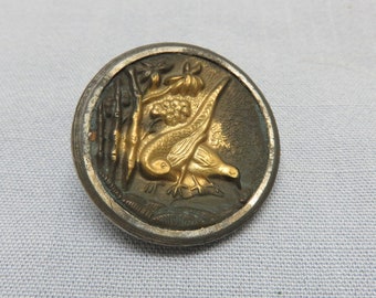 May include: A round, silver-toned button with a raised, gold-colored design of a bird perched on a branch. The button has a textured surface and a slightly raised rim.