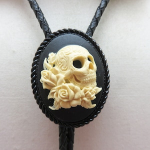 Pode incluir: Uma gravata bolo com um design de caveira e rosas creme sobre um fundo oval preto. A caveira é detalhada, e as rosas a rodeiam. A gravata tem um cordão trançado preto.