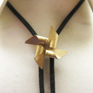 May include: A gold-colored metal pinwheel bolo tie with black braided cords. The pinwheel is a four-pointed star shape.