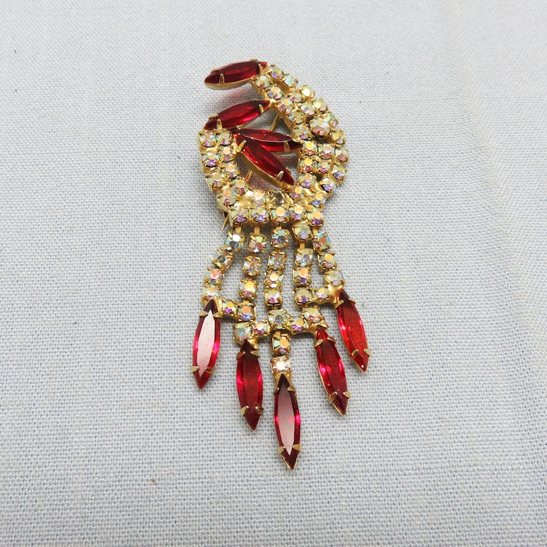 Pode incluir: Um broche dourado com um design pendente que apresenta strass vermelhos e transparentes. O broche tem um design de redemoinho na parte superior e uma franja de strass vermelhos na parte inferior.