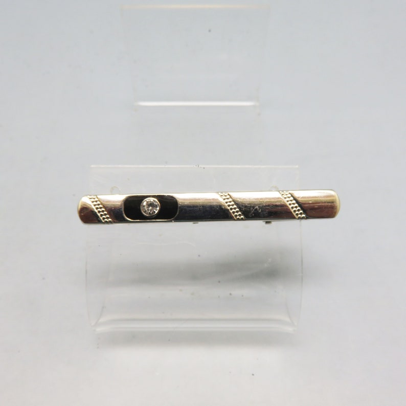 May include: Silver tie bar with a black enamel inlay and a single clear rhinestone in the center. The tie bar has a gold-toned design on the edges.