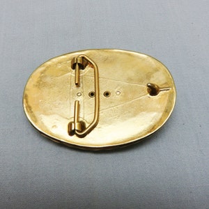 May include: A gold-toned oval belt buckle with a decorative design and a metal loop on the back.
