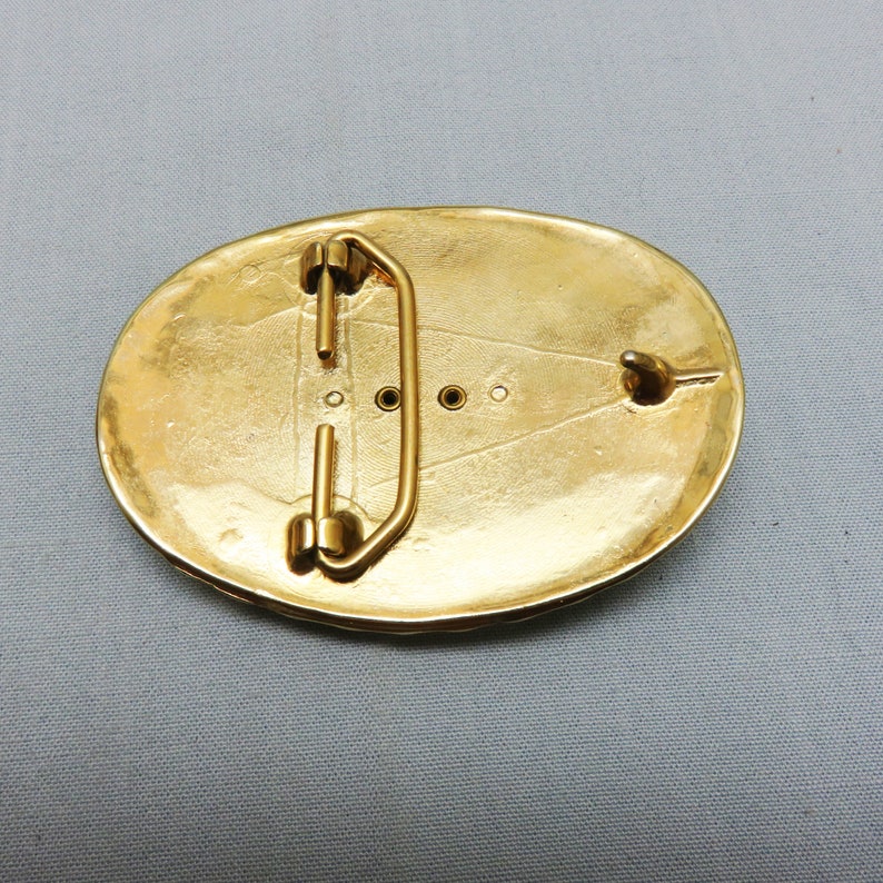 May include: A gold-toned oval belt buckle with a decorative design and a metal loop on the back.