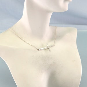 K&ouml;nnte beinhalten: Eine silberne Halskette mit einem zarten, geh&auml;mmerten Kreuzanh&auml;nger. Die Kette ist im einfachen, d&uuml;nnen Kabeldesign.