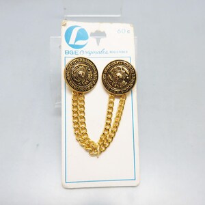 May include: Two gold-toned metal buttons connected by a double gold chain, mounted on a white BGE Originales display card. Each button features a circular design with embossed text reading NON CAPIT ORBIS LUDOVICUS REX.