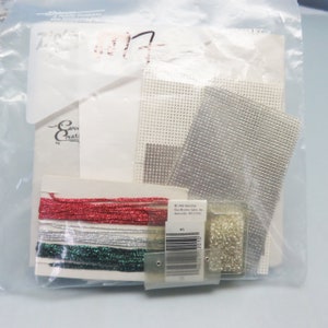 Op de afbeelding: Een plastic zak met een knutselset voor een kerstversiering. De set bevat wit Aida-doek, zilveren en witte kralen, rood, zilver en groen garen en een klein zakje met witte kralen.