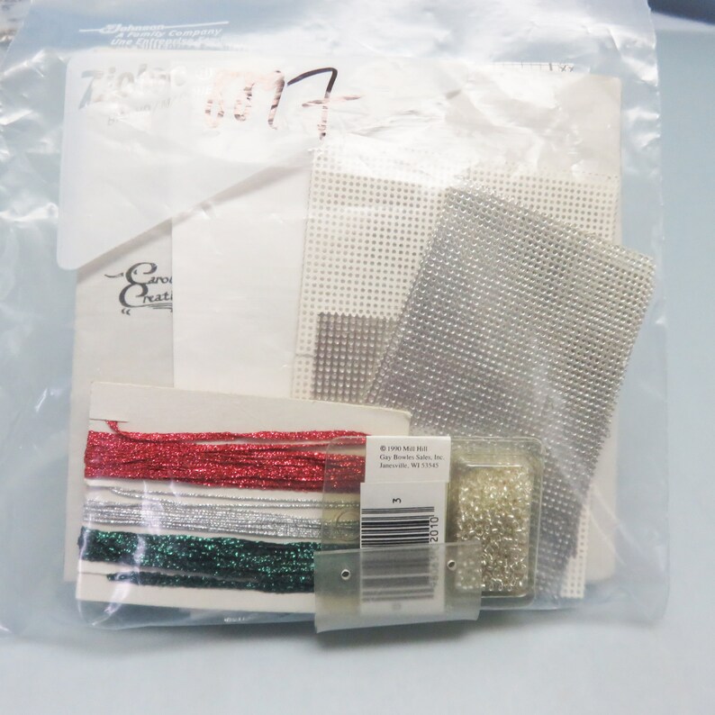 Op de afbeelding: Een plastic zak met een knutselset voor een kerstversiering. De set bevat wit Aida-doek, zilveren en witte kralen, rood, zilver en groen garen en een klein zakje met witte kralen.