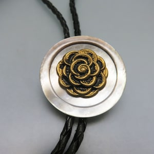 May include: A black leather bolo tie with a silver and gold round pendant featuring a black and gold rose design.