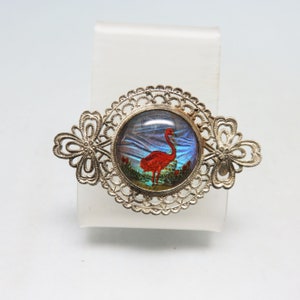 May include: A silver filigree brooch with a round glass cabochon featuring a red flamingo on a blue and green background.