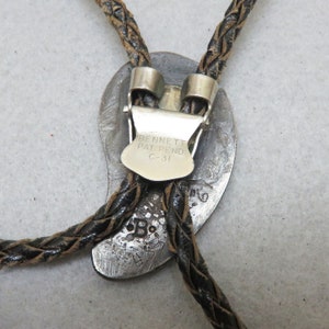 May include: Vintage bolo tie with a silver-toned oval pendant and braided leather cord. The pendant features the text "BENNETT PAT. PEND. C-31" and a stamped "B" marking. The cord is dark brown and the clasp is silver.