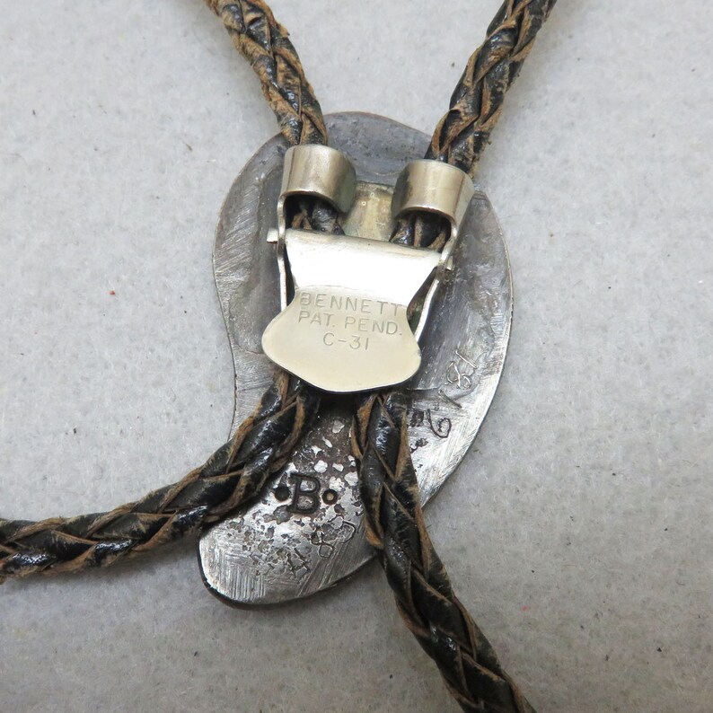 May include: Vintage bolo tie with a silver-toned oval pendant and braided leather cord. The pendant features the text "BENNETT PAT. PEND. C-31" and a stamped "B" marking. The cord is dark brown and the clasp is silver.