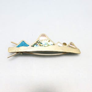 May include: A gold-toned metal hair clip featuring a mountain range design. The mountains are inlaid with turquoise, iridescent abalone shell, and mother-of-pearl. The clip has two prongs for securing it in the hair.