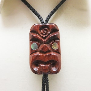 May include: A bolo tie featuring a rectangular, dark brown, hand-carved wooden pendant with a stylized face design. The pendant includes two circular, iridescent abalone shell inlays for eyes and is suspended from a black braided cord necklace.