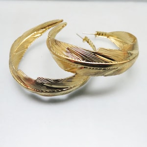 May include: A pair of gold hoop earrings with a feather design. The earrings are large and have a delicate, intricate design.