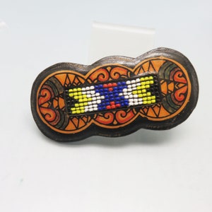 May include: A black leather hair clip with a colorful beaded design. The design features a geometric pattern with yellow, blue, white, red, and black beads.