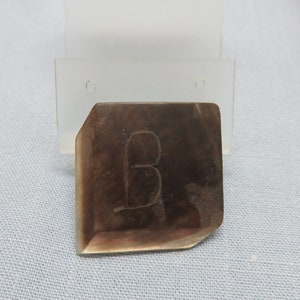 May include: A dark brown, rectangular, metal button with a carved letter 'B' on the front.
