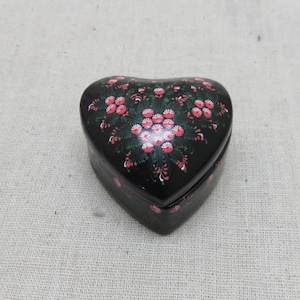 May include: A heart-shaped black box with a floral design. The box is painted with red and pink flowers and green leaves. The box is closed and sits on a neutral-coloured surface.