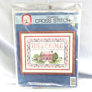 May include: A counted cross stitch pattern featuring a red house with a welcome sign, surrounded by pink hearts and green trees. The finished size is 12 inches by 12 inches and the matted size is 14 inches by 14 inches.