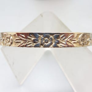 May include: A gold-toned bangle bracelet with a floral and leaf pattern. The detailed design features embossed flowers and foliage, creating a textured surface. The bracelet is displayed against a white background.