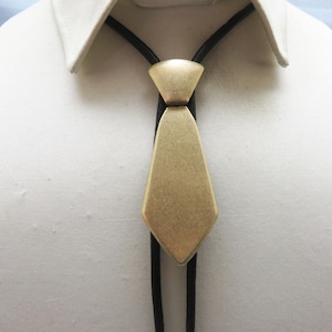 May include: A gold-colored bolo tie with a geometric design. The tie features a diamond-shaped pendant and a black cord. The bolo tie is worn against a white collared shirt.