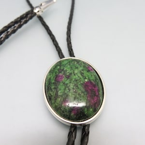 May include: A green and pink gemstone bolo tie with a silver setting. The bolo tie has a black braided cord.