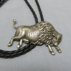 May include: A silver bolo tie featuring a detailed, three-dimensional bison design. The bolo tie is on a black braided cord.