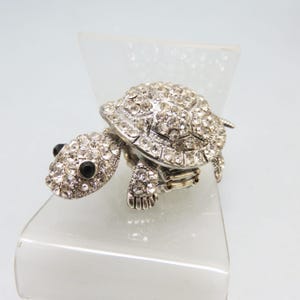 May include: A silver-toned turtle figurine encrusted with clear rhinestones. The turtle's shell, head, and legs are covered in sparkling crystals. The eyes are black. The figurine is displayed on a clear, angled stand.