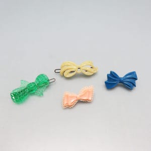 May include: Four colorful hair bows in a variety of styles. The bows are in shades of green, yellow, peach, and blue. The green bow is a clip-on style, while the yellow bow is a barrette. The peach bow has a lace trim, and the blue bow is a simple design.