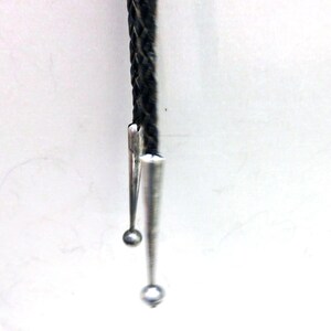 May include: A close-up of a bolo tie. The tie features a braided black cord with silver-colored metal tips and a decorative slide. The tips are cone-shaped with small, round accents at the ends. The background is plain white.