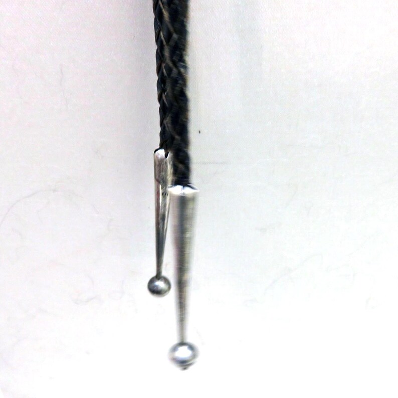 May include: A close-up of a bolo tie. The tie features a braided black cord with silver-colored metal tips and a decorative slide. The tips are cone-shaped with small, round accents at the ends. The background is plain white.