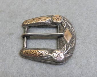 May include: Silver-tone belt buckle with gold-tone floral and leaf accents. The buckle features a diamond-shaped gold-tone center and a silver bar for the belt. The design includes detailed floral and leaf patterns.