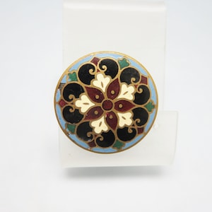 May include: A round, vintage brooch with a colorful floral design. The brooch features a gold-toned metal frame and a central flower with burgundy, cream, and black petals. A light blue border surrounds the design, with green accents.