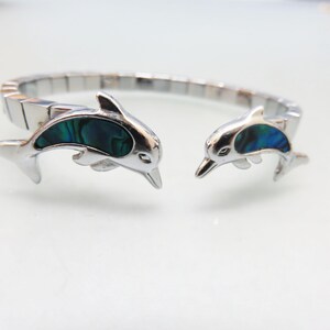 May include: Silver-toned bracelet featuring two dolphin charms. Each dolphin has a blue and green iridescent inlay. The bracelet is made of rectangular links and is designed to open and close.