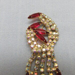 Pode incluir: Um broche dourado com um design em cascata de strass transparentes e iridescentes. A parte superior do broche apresenta um grupo de pedras vermelhas facetadas.