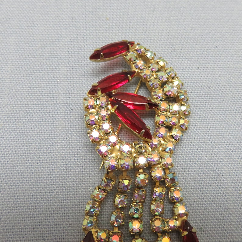 Pode incluir: Um broche dourado com um design em cascata de strass transparentes e iridescentes. A parte superior do broche apresenta um grupo de pedras vermelhas facetadas.
