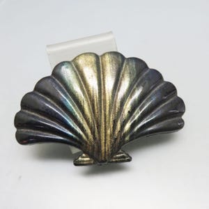 May include: A silver-toned seashell-shaped brooch with a fan-like design. The brooch has a metallic sheen with shades of gold, blue, and silver. The seashell design is detailed with ridges and curves.