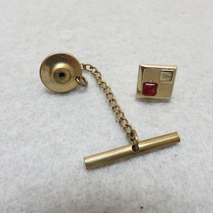 May include: Gold tie tack with a red and white square design. The tie tack has a chain and a pin.