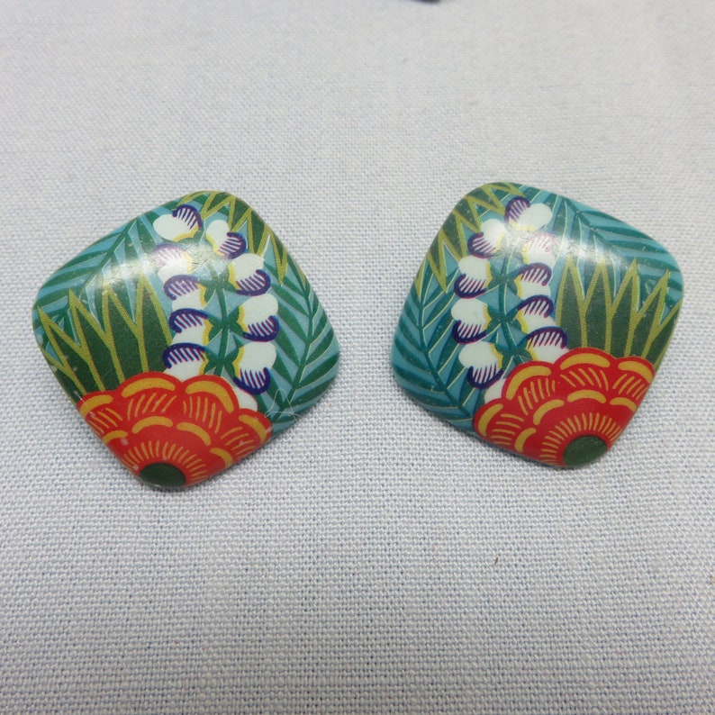 Op de afbeelding: Een paar vierkante oorbellen met een bloemenmotief. De oorbellen zijn turquoiseblauw met groene bladeren, witte bloemen en oranje en rode accenten.