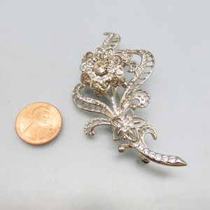 May include: Silver tone brooch with a floral design featuring a large flower with rhinestones and smaller flowers with rhinestones. The brooch is about the size of a US penny.