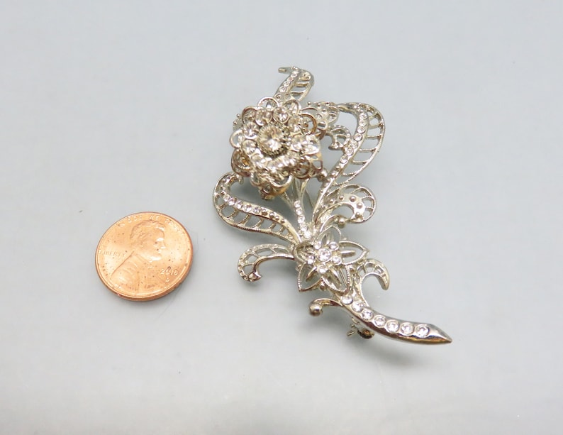 May include: Silver tone brooch with a floral design featuring a large flower with rhinestones and smaller flowers with rhinestones. The brooch is about the size of a US penny.