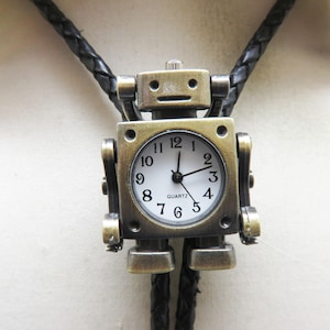 May include: A bronze-colored robot-shaped bolo tie with a white clock face. The clock has black hands and the word "QUARTZ" at the bottom. The tie has a black braided leather cord.