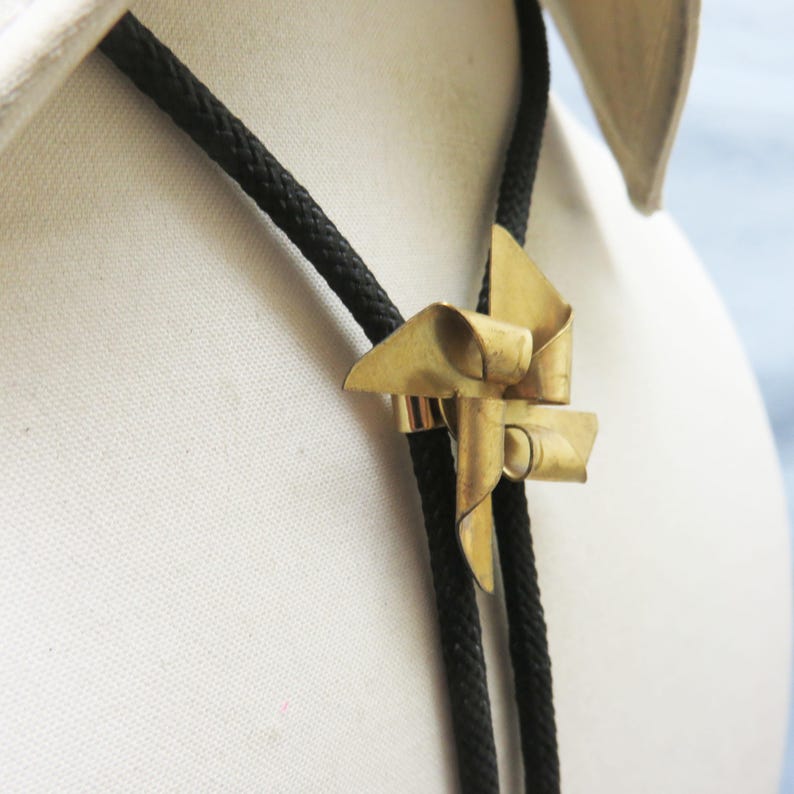 May include: A gold-colored metal bolo tie with a bow-shaped design. The tie is on a black cord and is worn on a white mannequin.